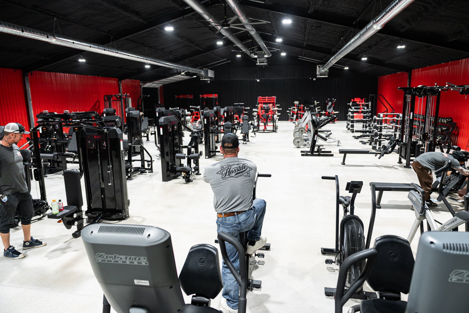 A finished gym floor of remanufactured cardio and strength machines with three people setting up and training, a red accent wall behind them.